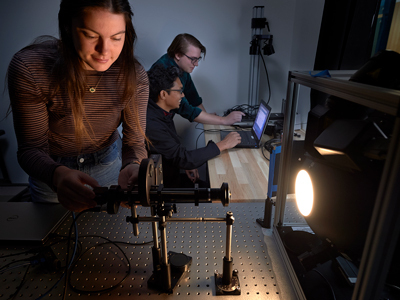 A person with long dark hair touching a piece of equipment that's shining a light at a surface. There are two people at a laptop in the background